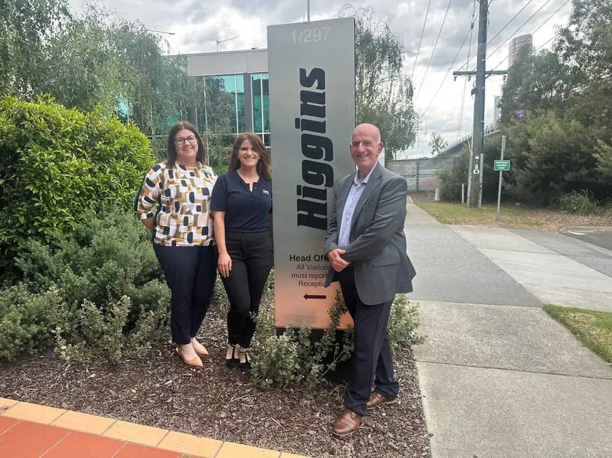 Two women and one man standing outdoors beside the Higgins Head Office sign.