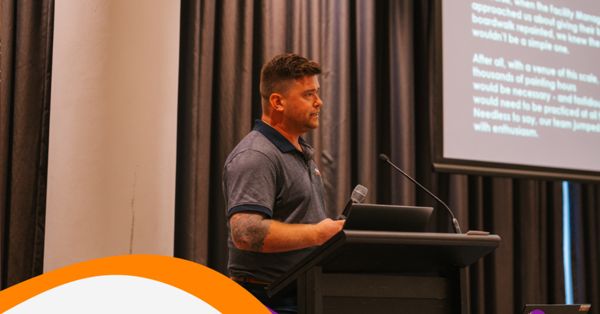 Brendan Childs in a grey polo shirt, speaking into a microphone at a lectern during a presentation.