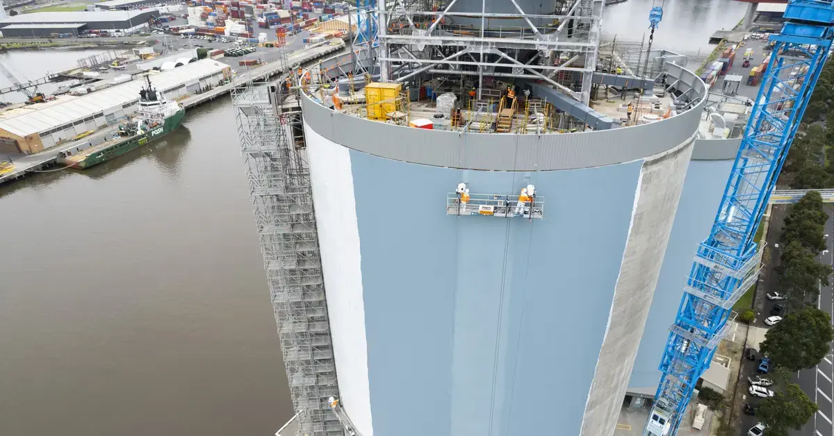 Aerial view of two workers on a swing stage painting a large industrial silo light blue.
