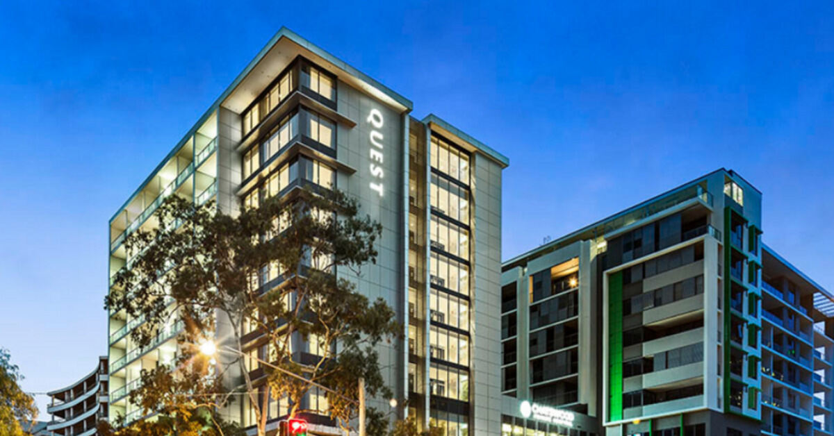 The modern Quest apartment hotel building on a city street corner at dusk.
