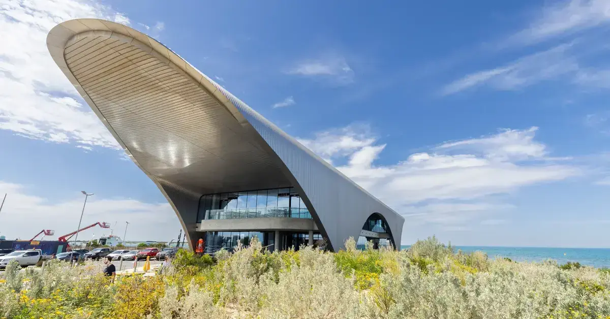 A modern building with a large, curved metal roof next to sand dunes and the ocean.
