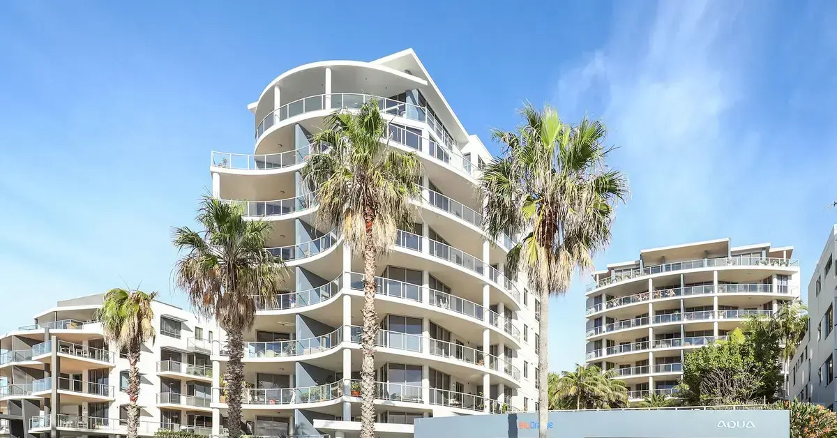Street view of modern, white apartment buildings with curved balconies and palm trees.