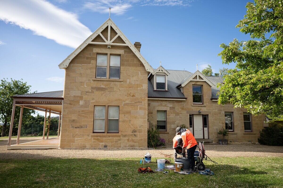 A worker in an orange vest prepares painting equipment on the lawn in front of a large stone house.