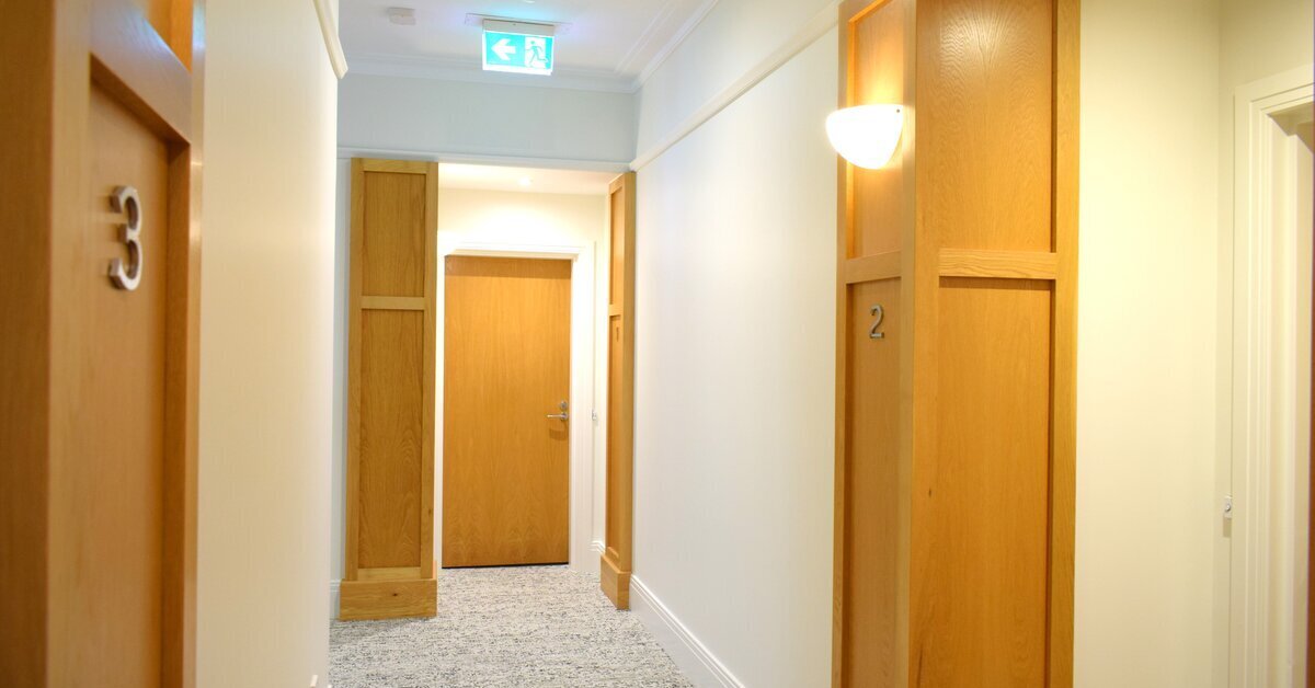 A building interior hallway with white walls and light wood trim around the doors.
