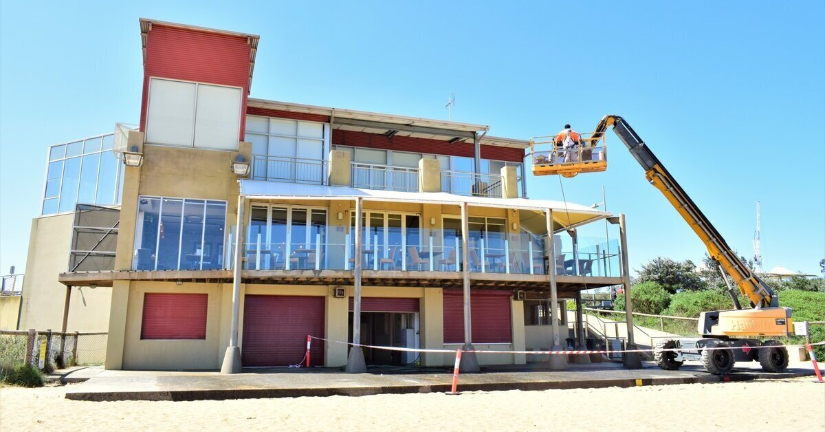 Two workers in a boom lift painting the exterior of a multi-storey beachside building.