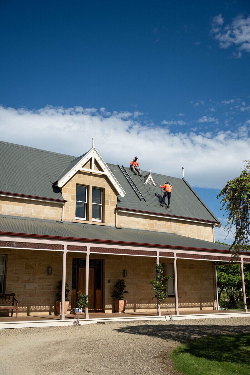 Two workers in orange vests use a ladder on the grey roof of a large stone house.